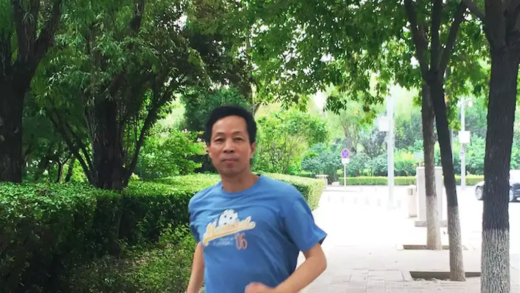 Hua-Yang - A man in a blue t-shirt and gray shorts is jogging on a tree-lined sidewalk, wearing neon running shoes, with greenery in the background.