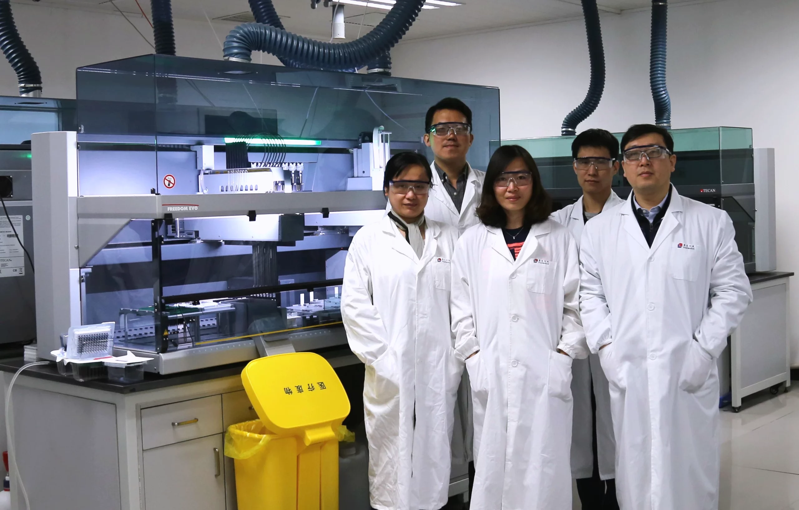 Fang and her team of scientists in lab coats pose in front of an advanced lab automation machine, with a biohazard bin in the foreground.
