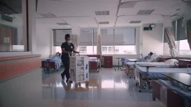 A healthcare professional pushes a medical cart in a clinical research ward, while a patient rests in bed, indicating early phase clinical research.
