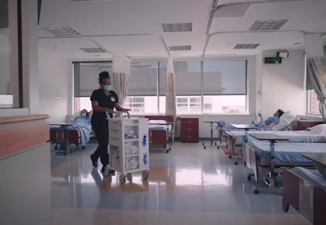 A healthcare professional pushes a medical cart in a clinical research ward, while a patient rests in bed, indicating early phase clinical research.