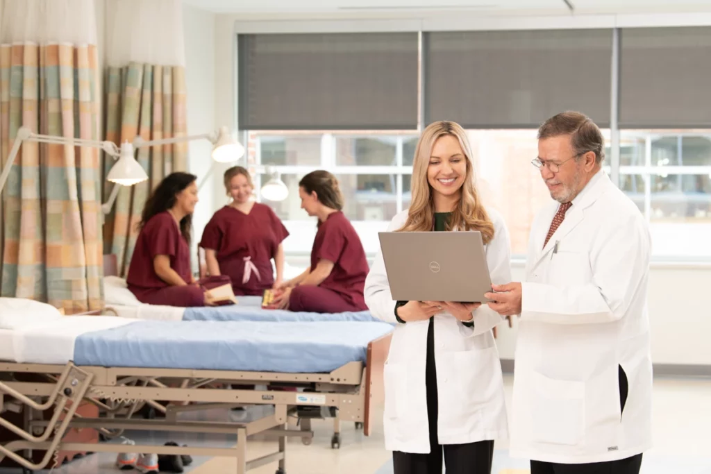 Scientists in lab coats review data on a laptop while clinical staff in scrubs interact with patients in a Baltimore clinic setting.