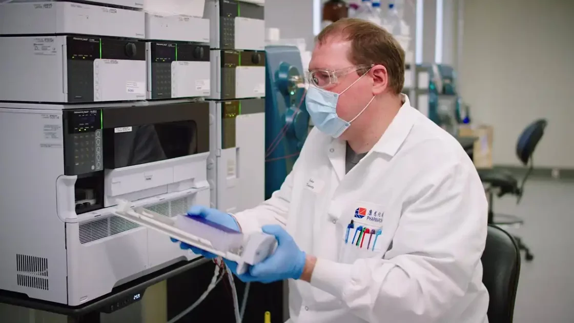 A scientist in a lab coat, safety glasses, and a face mask operates analytical equipment in a laboratory. Surrounded by various scientific instruments and machines, they handle a tray with samples for preclinical ADME studies in an environment that is clean and organized.