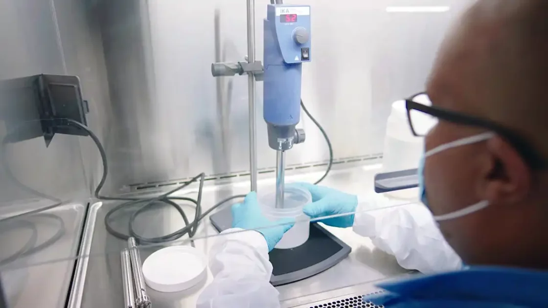A person wearing a face mask, gloves, and protective clothing is operating laboratory equipment. They are working under a fume hood with a blue machine, possibly mixing or conducting an experiment related to genetic toxicology. Various lab tools and containers are on the stainless steel surface.