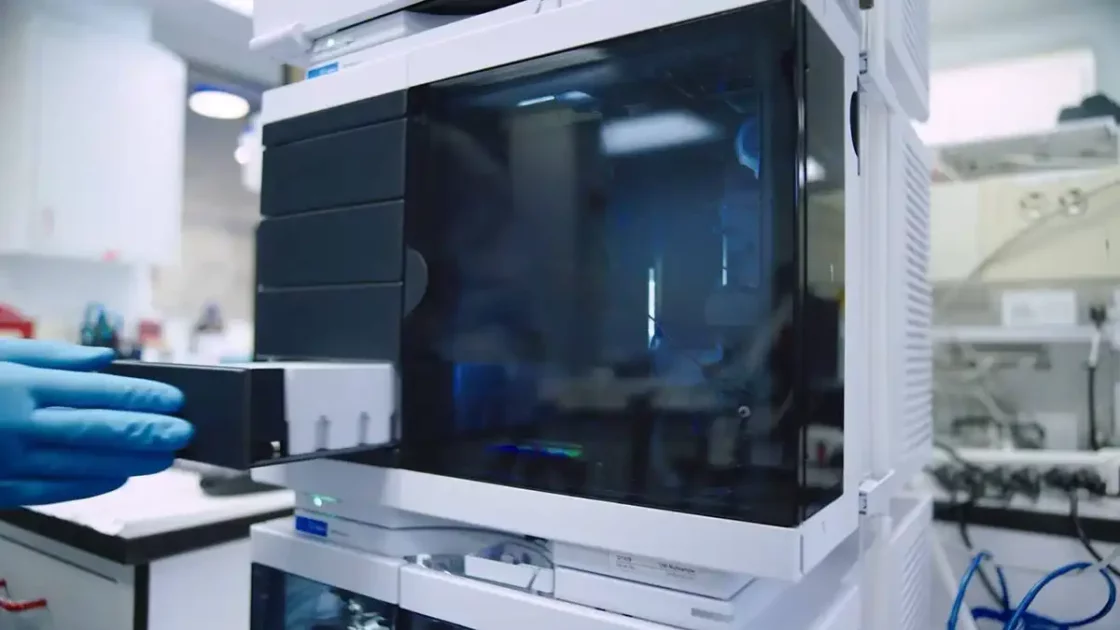 A scientist wearing blue gloves loads a sample into an Agilent HPLC system, featuring a black-paneled autosampler and stacked analytical modules in a laboratory setting.
