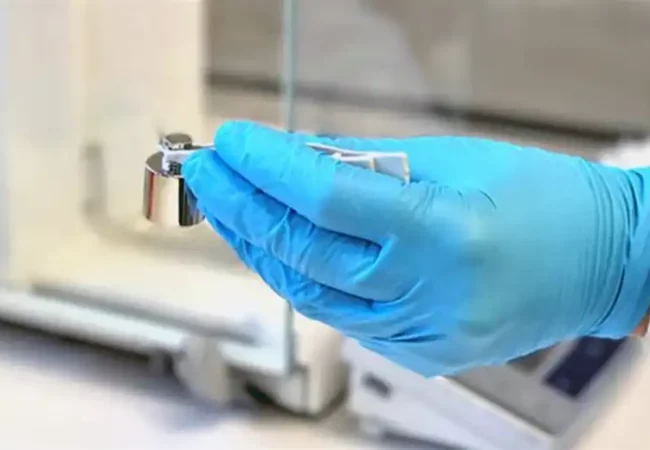 A scientist wearing a blue glove carefully handles a small calibration weight with tweezers, likely for precision measurement in a microdosing study.