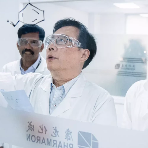 Scientist in safety goggles analyzing a chemical structure on a glass panel, collaborating with colleagues in a laboratory setting with Pharmaron branding.