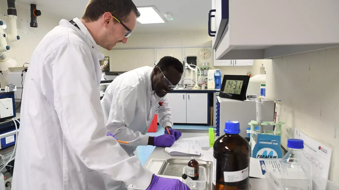 Two scientists in a laboratory work together, reviewing notes and handling chemicals, with bottles and equipment labeled for a Carbon-14 clinical program.