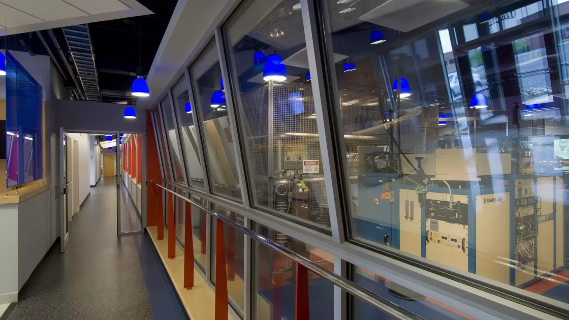 A modern laboratory hallway with large windows showcasing an Accelerator Mass Spectrometry (AMS) system, illuminated by blue pendant lights.