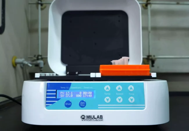 Laboratory equipment with an open lid displays a digital interface showing temperature, speed, and time settings. Orange tube holders and test tubes used in antibody-drug conjugates research are placed inside.