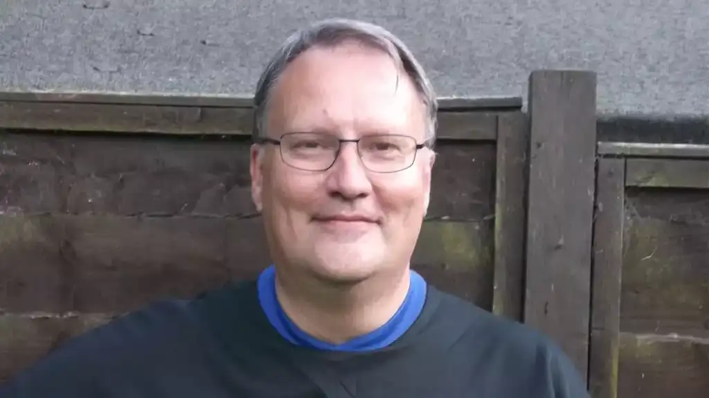 A man wearing glasses and a black football jersey with the "Hotpoint" logo, holding a soccer ball, stands in front of a wooden fence.