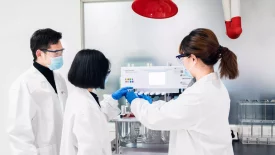 Three scientists wearing lab coats and masks operate an analytical instrument for quality control, reviewing data and processes in a sterile lab environment.