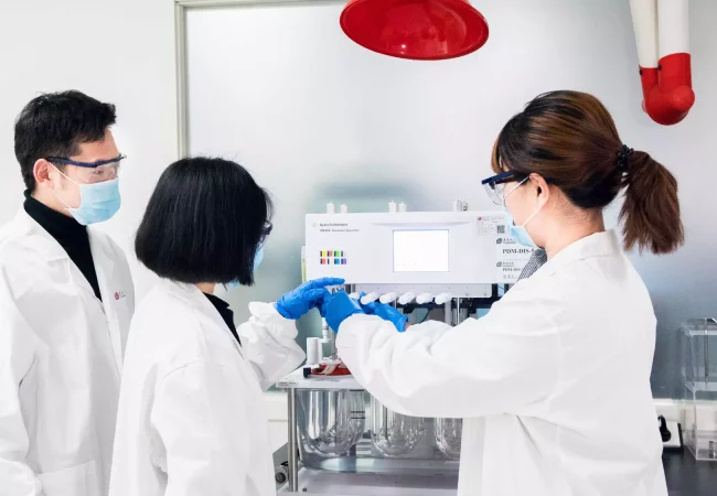 Three scientists wearing lab coats and masks operate an analytical instrument for quality control, reviewing data and processes in a sterile lab environment.
