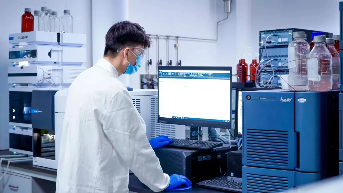 A scientist wearing protective gear operates a computer connected to advanced laboratory equipment, analyzing samples in a quality control lab environment.