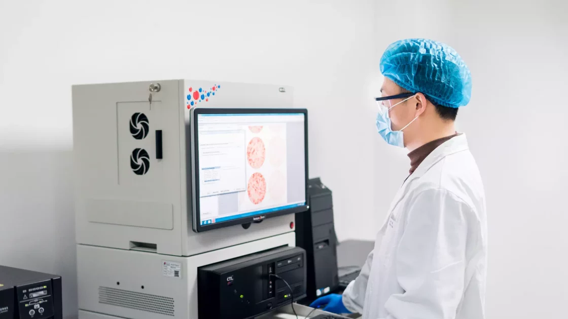A scientist in a lab coat, mask, and hair cover analyzes immunology data from a digital monitor, related to animal disease models and biomarker studies.