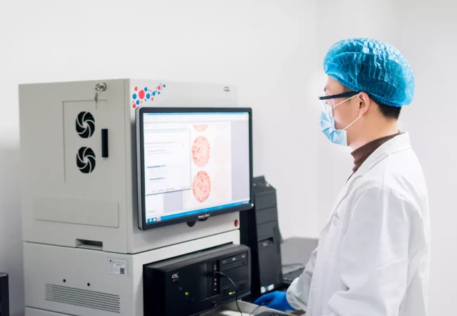 A scientist in a lab coat, mask, and hair cover analyzes immunology data from a digital monitor, related to animal disease models and biomarker studies.
