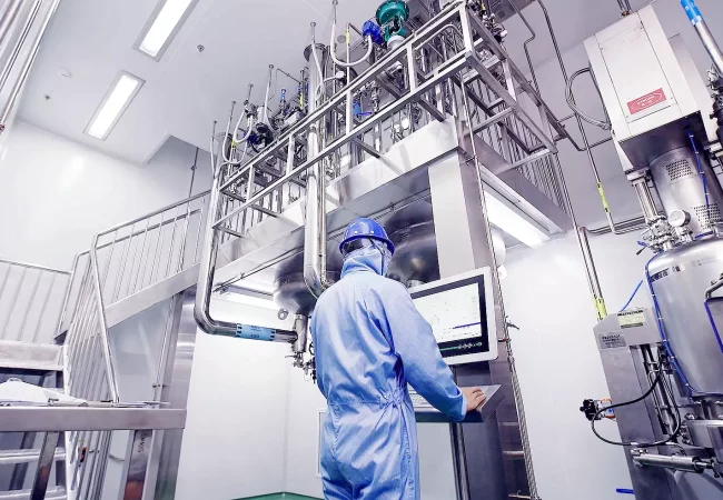A technician in full protective gear monitors equipment through a control screen in a high-tech API (Active Pharmaceutical Ingredient) manufacturing facility.
