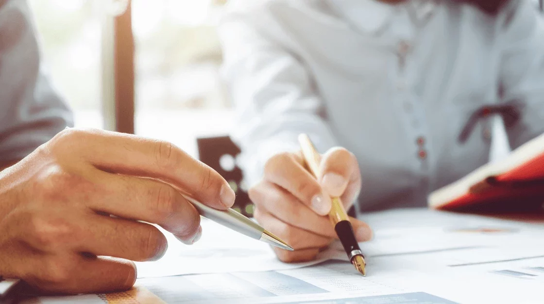 Two scientists review documents with pens in hand, focusing on data for quality assurance audit purposes. A red notebook is visible in the background.