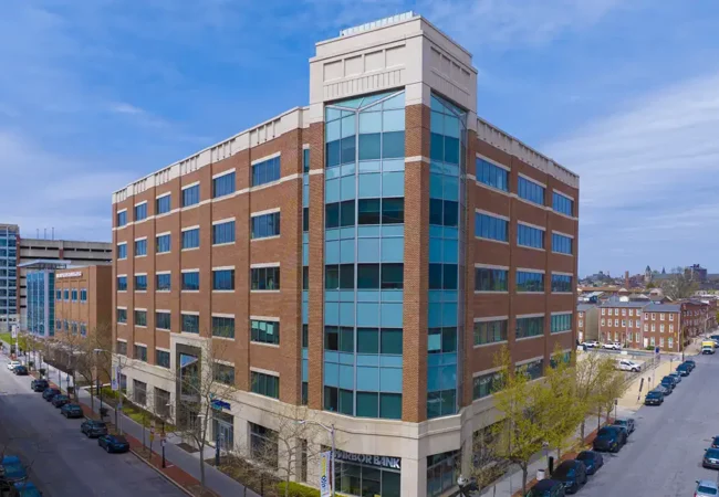 A multi-story brick and glass office building located in Baltimore, with the Pharmaron logo displayed, surrounded by trees and street is the Baltimore Clinical Pharmacology Center