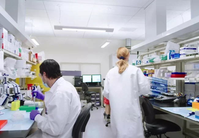 Scientists in lab coats work at a well-equipped laboratory, handling pipettes and conducting experiments related to vaccine development.