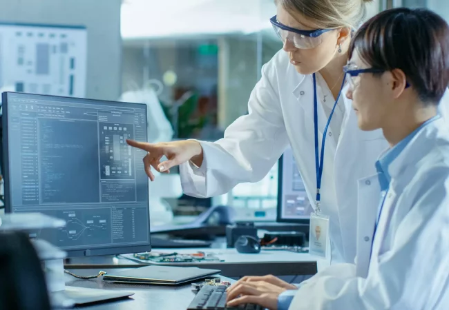 Two scientists in lab coats and safety glasses reviewing data on a computer screen, discussing biometric analysis in a research environment.