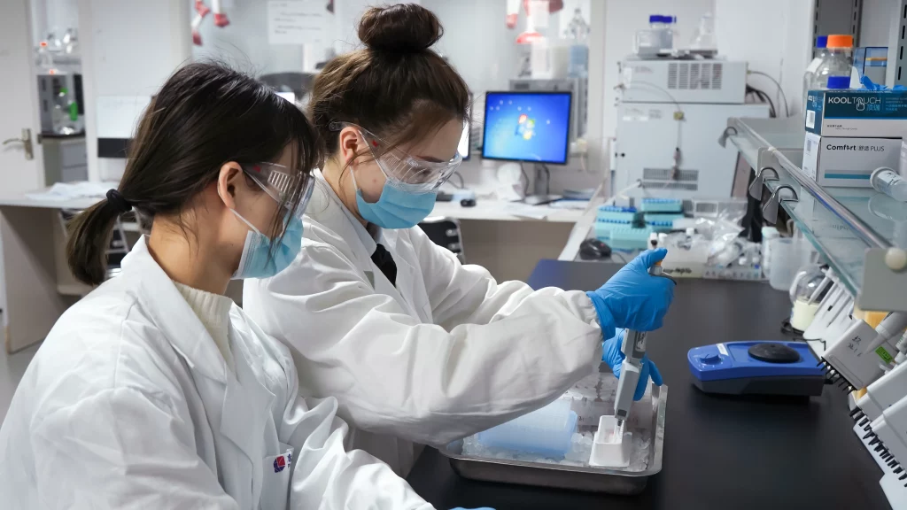 Two scientists in lab coats and masks conduct an experiment using a pipette in a biosciences lab, with equipment and supplies on the counter.