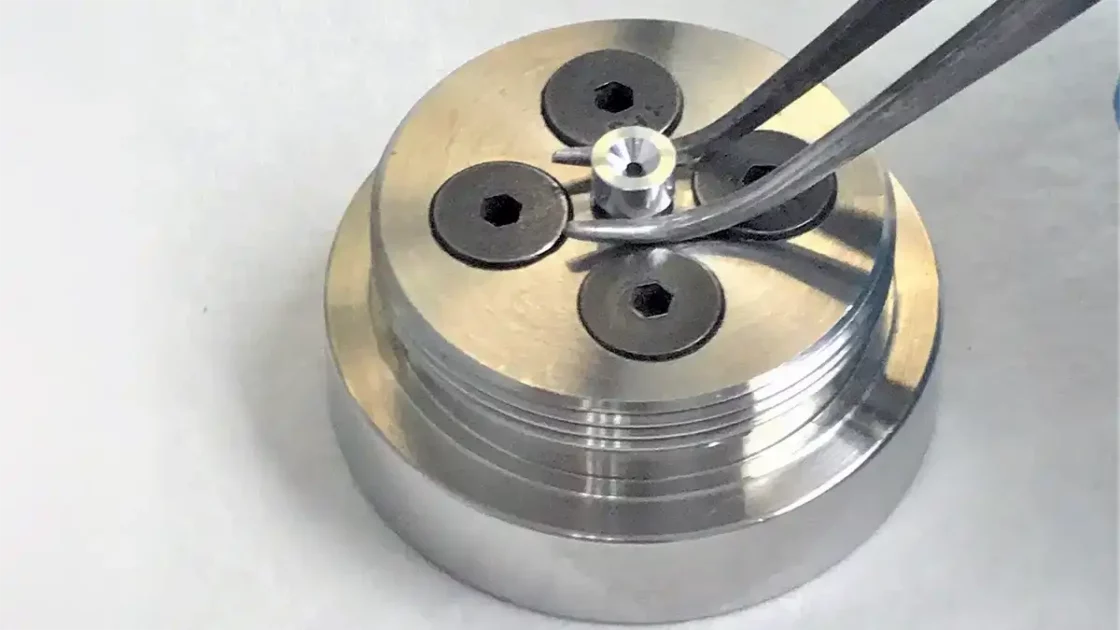 A scientist wearing a blue glove uses tweezers to handle a small component on a metallic Carbon-14 Microtracer cathode with hexagonal screws.