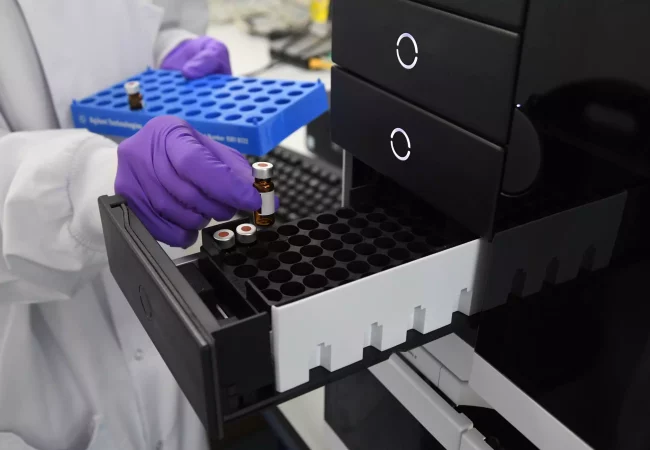 A scientist places vials into a high-capacity tray, preparing samples for analysis in a Carbon-14 radiolabeling and AMS system for biologics research.