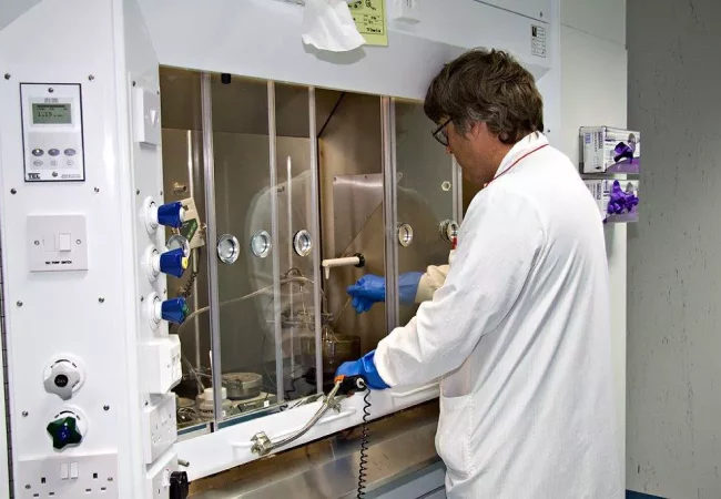 A scientist works in an enclosed fume hood conducting Carbon-14 radiosynthesis, wearing protective gloves and handling specialized equipment.
