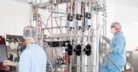 Two scientists in protective gear work in a CDMO plasmid manufacturing suite. One operates a control panel, while the other adjusts equipment on a large, stainless steel bioreactor system, designed for producing plasmid DNA at scale. Tubing, valves, and sensors emphasize precise, high-tech biopharmaceutical production.
