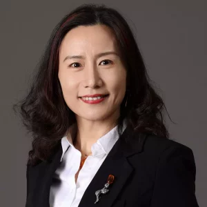 Celina Zhong, MBA - A woman with long, dark hair is smiling. She is wearing a black blazer over a white shirt. The blazer has two decorative pins on the lapel – one shaped like a red ribbon and the other resembling a figure, symbolizing her expertise in bioanalytical development. The background is a solid, dark gray color.