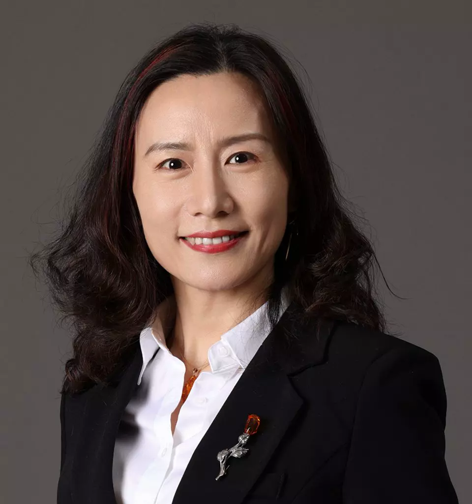 Celina Zhong, MBA - A woman with long, dark hair is smiling. She is wearing a black blazer over a white shirt. The blazer has two decorative pins on the lapel – one shaped like a red ribbon and the other resembling a figure, symbolizing her expertise in bioanalytical development. The background is a solid, dark gray color.
