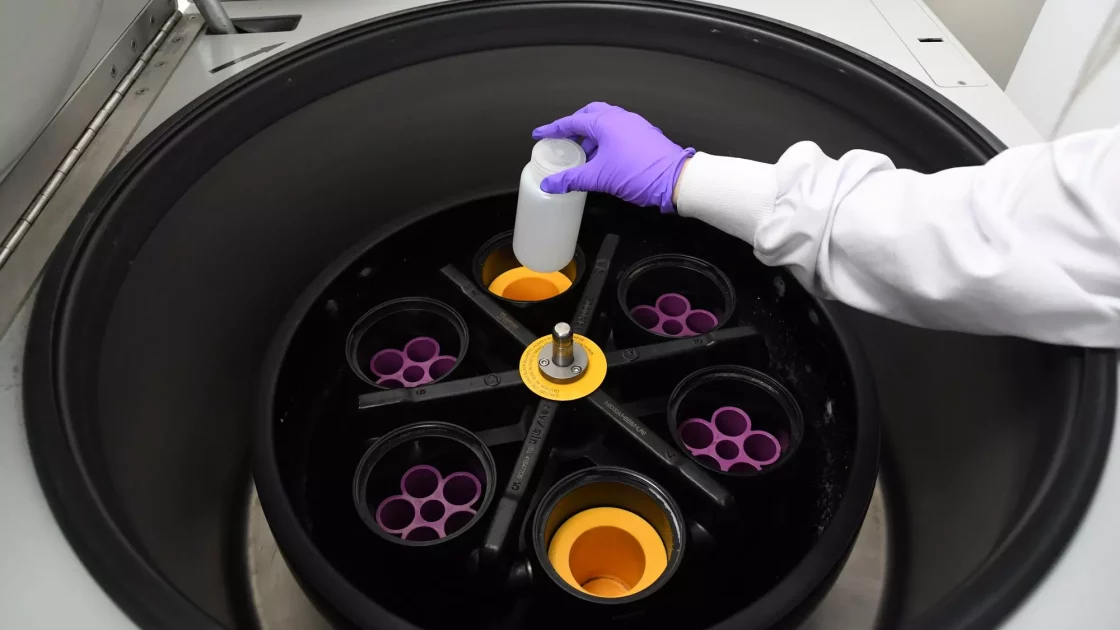 A scientist places a container inside a centrifuge rotor with labeled slots, preparing samples for analysis in environmental metabolism research.