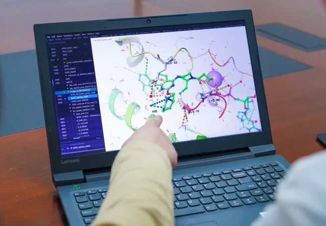 Two scientists review a 3D molecular model on a laptop, discussing computer-aided drug design. One points to specific binding sites, while a sidebar displays structural data and analysis.