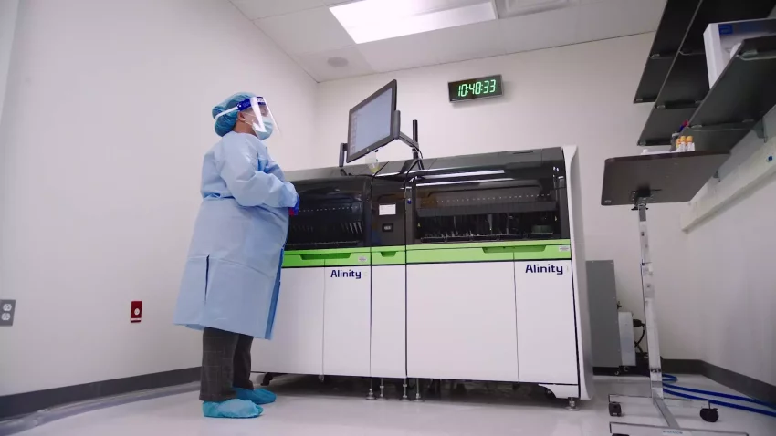 A scientist in full protective gear operates an Alinity diagnostic machine in a clinical development laboratory, closely monitoring the screen.