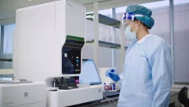 A scientist in protective gear operates a machine in a lab during vaccine development and infectious challenge studies, monitoring data on a nearby screen.