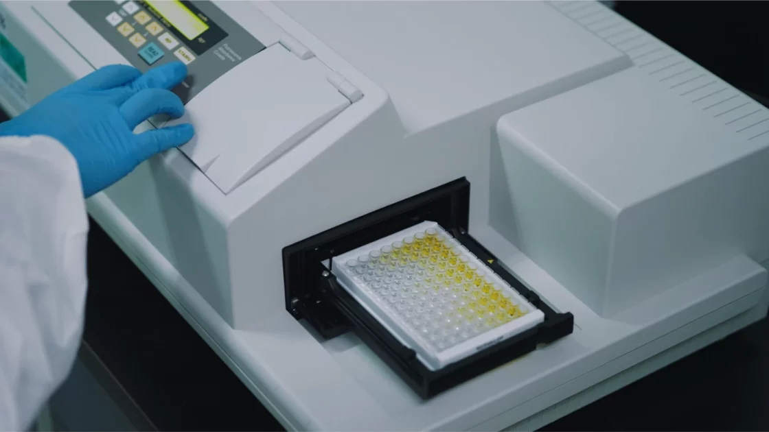 A scientist's gloved hand operates a lab instrument analyzing a multi-well plate with yellow samples, used for developmental and reproductive toxicology testing.