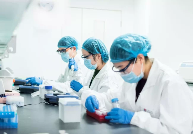 Three scientists wearing lab coats, masks, and hairnets work with pipettes and samples in a laboratory, focused on discovery DMPK studies.