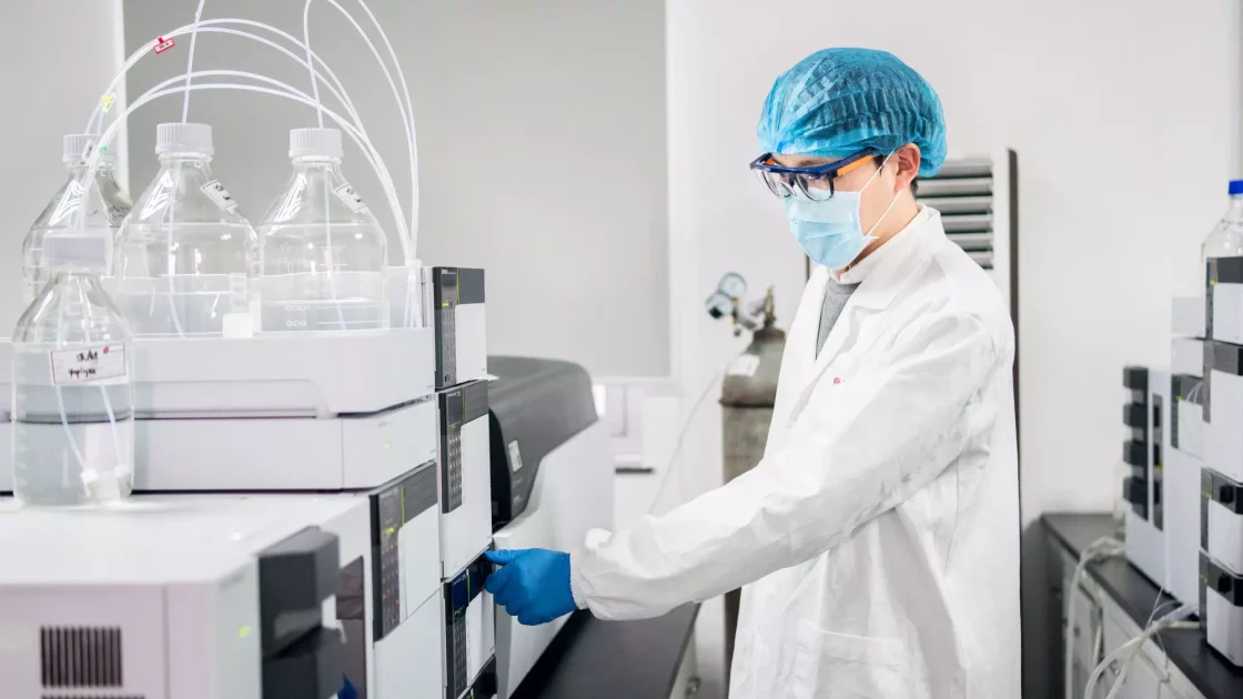 A scientist wearing a white lab coat, blue hair net, safety glasses, and a face mask operates laboratory equipment with multiple bottles of liquids. The sterile and modern lab setting is equipped for advanced preclinical ADME studies, with various scientific devices and containers visible.