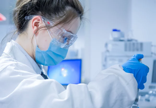 A scientist in protective gear uses a pipette in a laboratory, performing Discovery in vivo pharmacokinetics (PK) research with analytical equipment in the background.