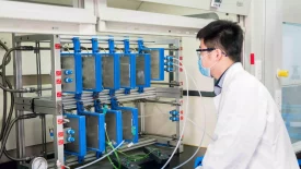 A scientist wearing protective gear operates a process chemistry apparatus with multiple blue reaction chambers and tubing connections in a lab setting.