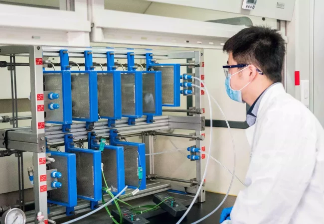 A scientist wearing protective gear operates a process chemistry apparatus with multiple blue reaction chambers and tubing connections in a lab setting.