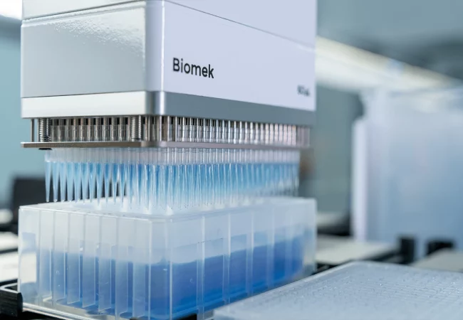 Close-up of a Biomek automated liquid handler with multiple pipettes dispensing blue liquid into a well plate for DMPK studies.