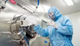 A scientist in full protective gear operates specialized equipment in a sterile environment for drug product manufacturing.