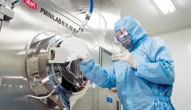 A scientist in full protective gear operates specialized equipment in a sterile environment for drug product manufacturing.