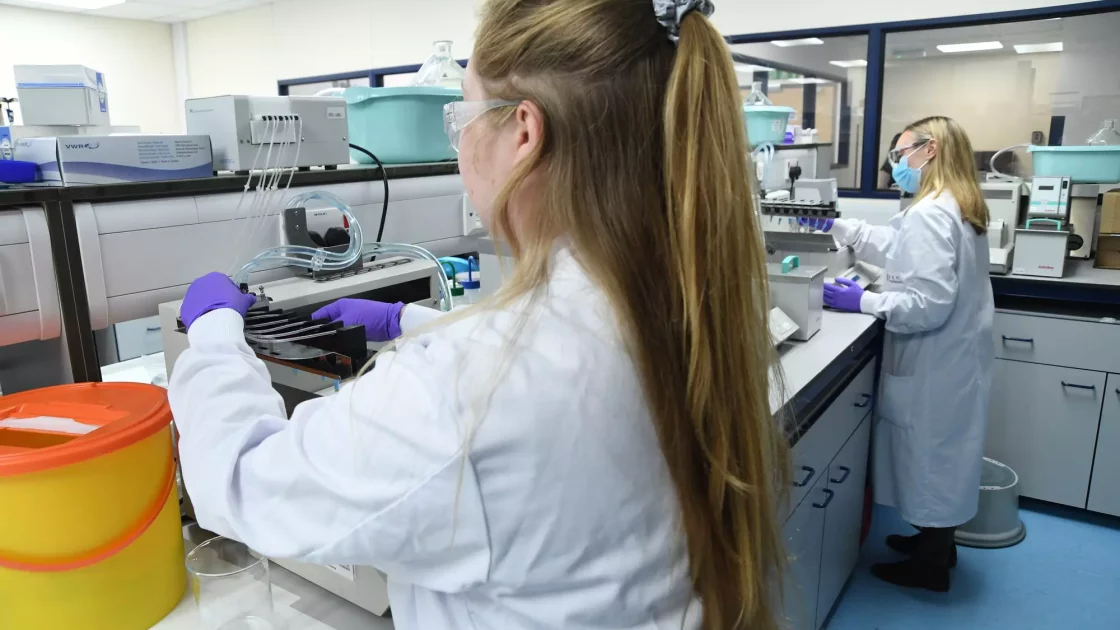 Scientists in a lab working with specialized equipment, conducting environmental metabolism studies. Both wear protective lab coats and gloves.