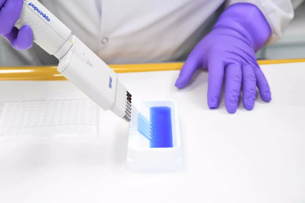 A scientist in purple gloves uses a multi-channel pipette to transfer a blue liquid into a well plate, possibly for integrated radiolabeled sciences research. The setup suggests precision handling of radioactive compounds for advanced assays.