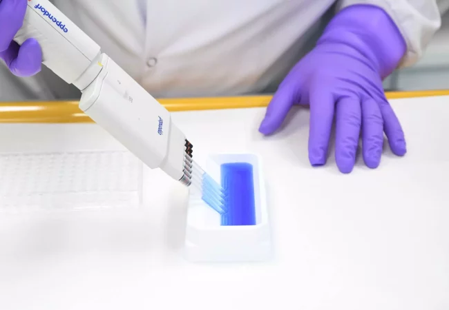 A scientist in purple gloves uses a multi-channel pipette to transfer a blue liquid into a well plate, possibly for integrated radiolabeled sciences research. The setup suggests precision handling of radioactive compounds for advanced assays.