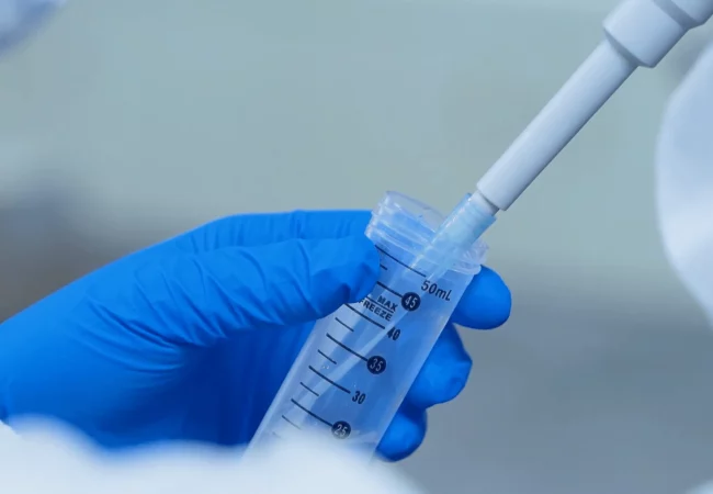 Close-up of a person wearing blue gloves and a white lab coat using a pipette to transfer a clear liquid into a graduated test tube marked up to 50 mL. The background is blurred, focusing attention on the laboratory equipment used in genetic toxicology research.
