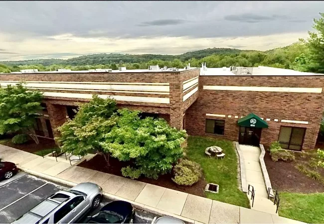 A low-rise brick building surrounded by trees and parking, featuring an entrance with a green canopy labeled "600," located at the Exton Pharmaron site.