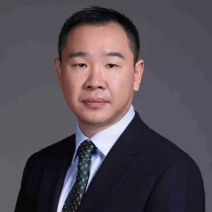 Gilbert Li, CFA, CPA - A man in a dark suit and a patterned tie with a light blue shirt poses against a plain gray background. With short black hair and a neutral expression, he exudes the precision of bioanalytical development.